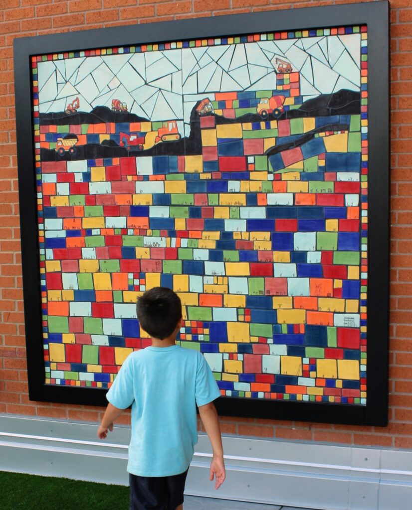 A child in a blue t-shirt stands in front of a colorful tile mosaic. The tiles connect to create a scene of construction workers with heavy duty equipment. The mosaic has a black frame and sits on a red brick wall.